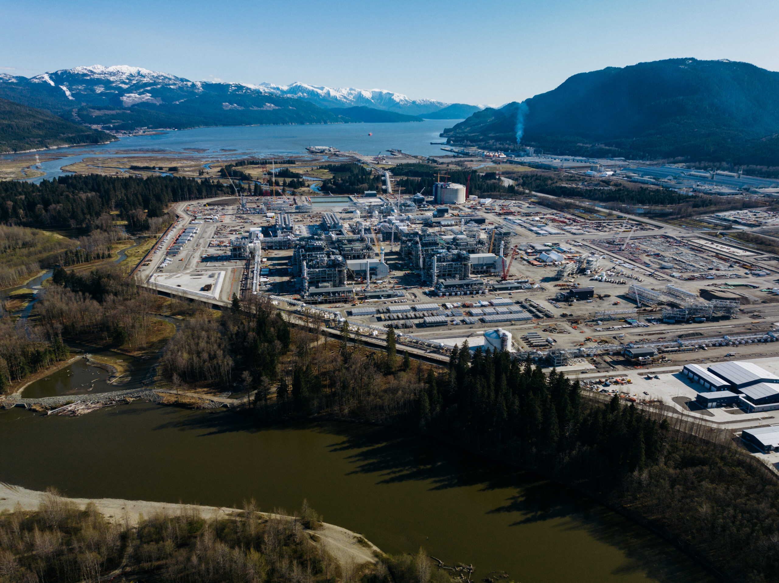 An aerial view of the LNG Canada liquified natural gas production facility add export terminal with the inlet in the background and snow capped mountains beyond