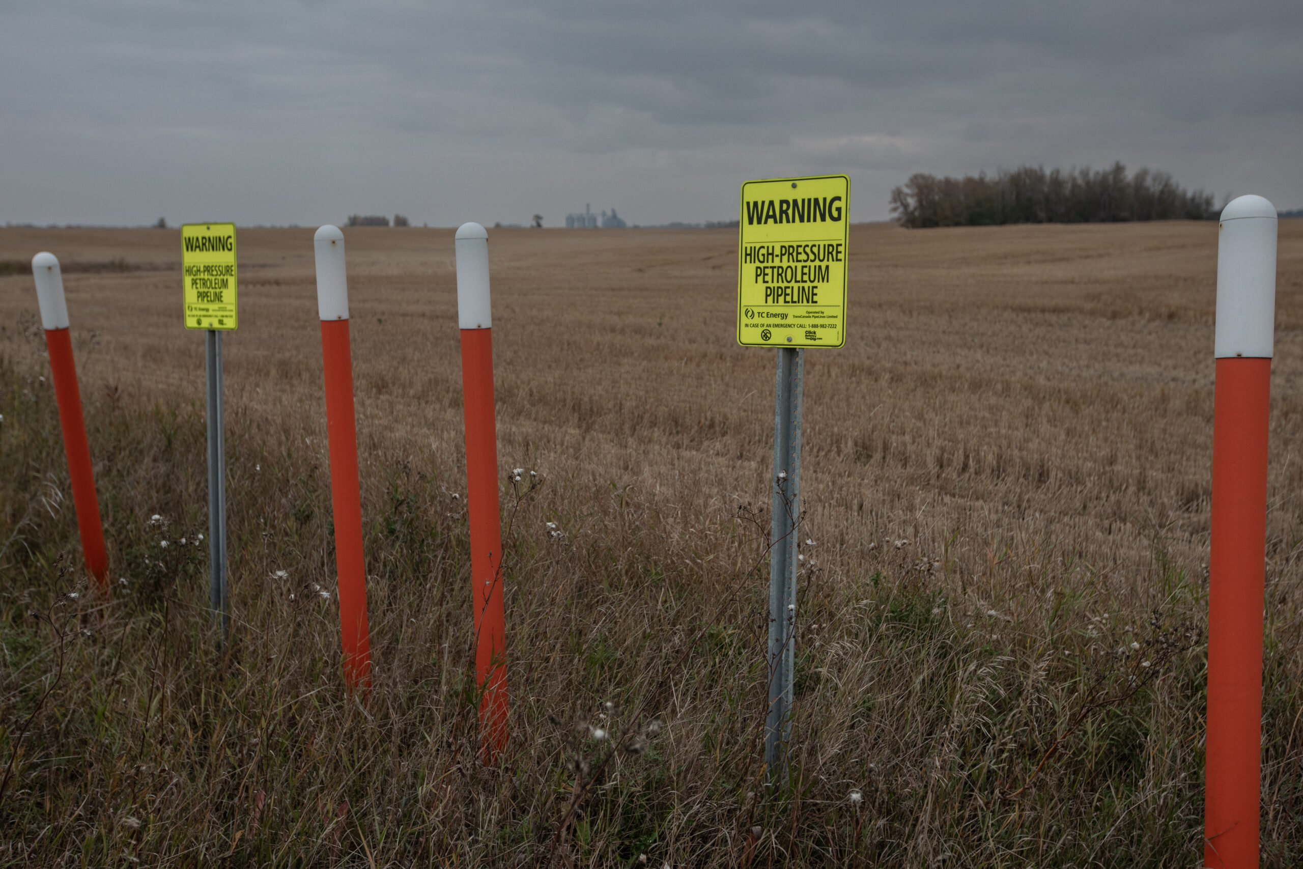Yellow sign that reads "warning: high pressure petroleum pipeline" in front of a field with a farm in the background