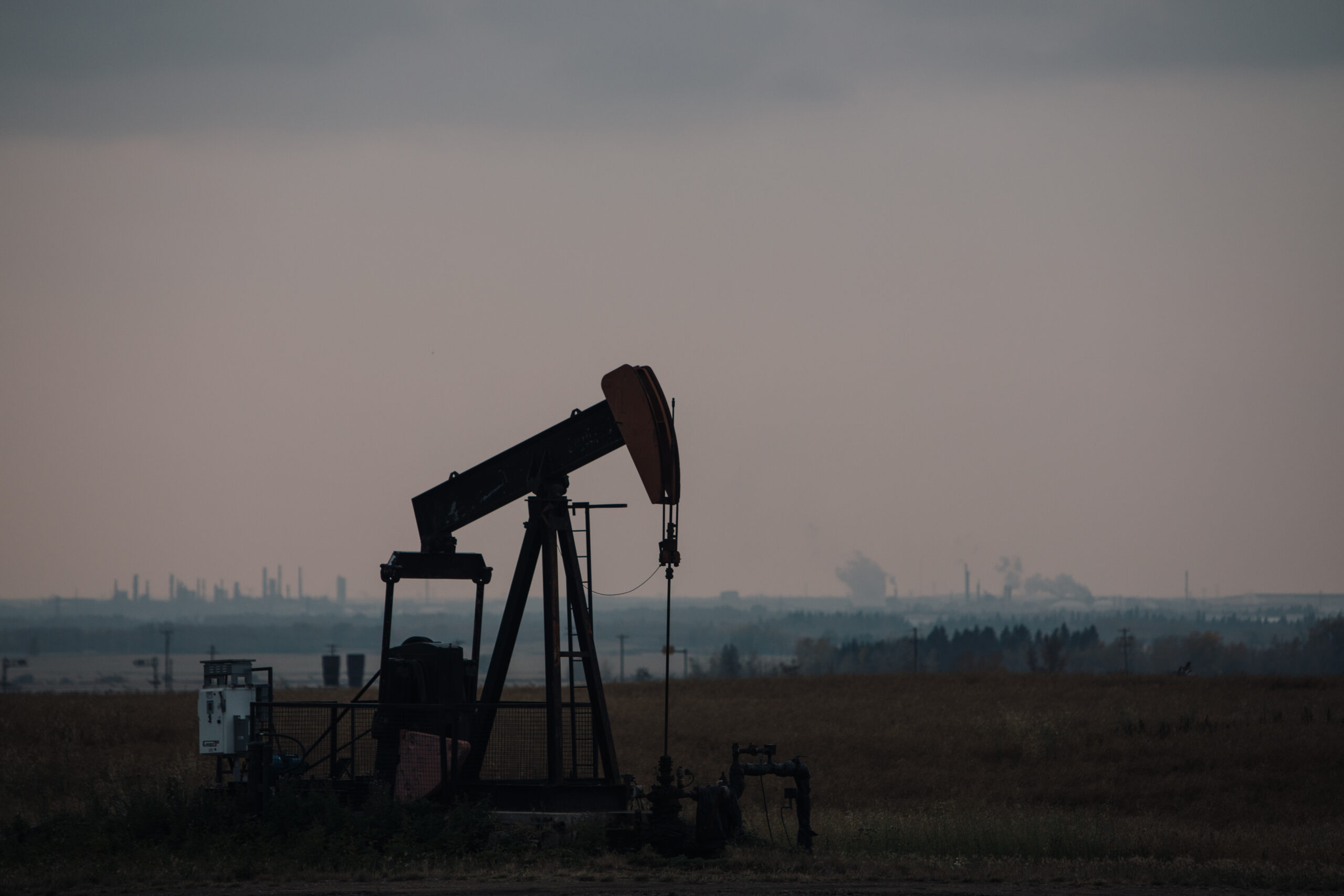 Pumpjack set against a dark grey sky with industrial activity in the distance.