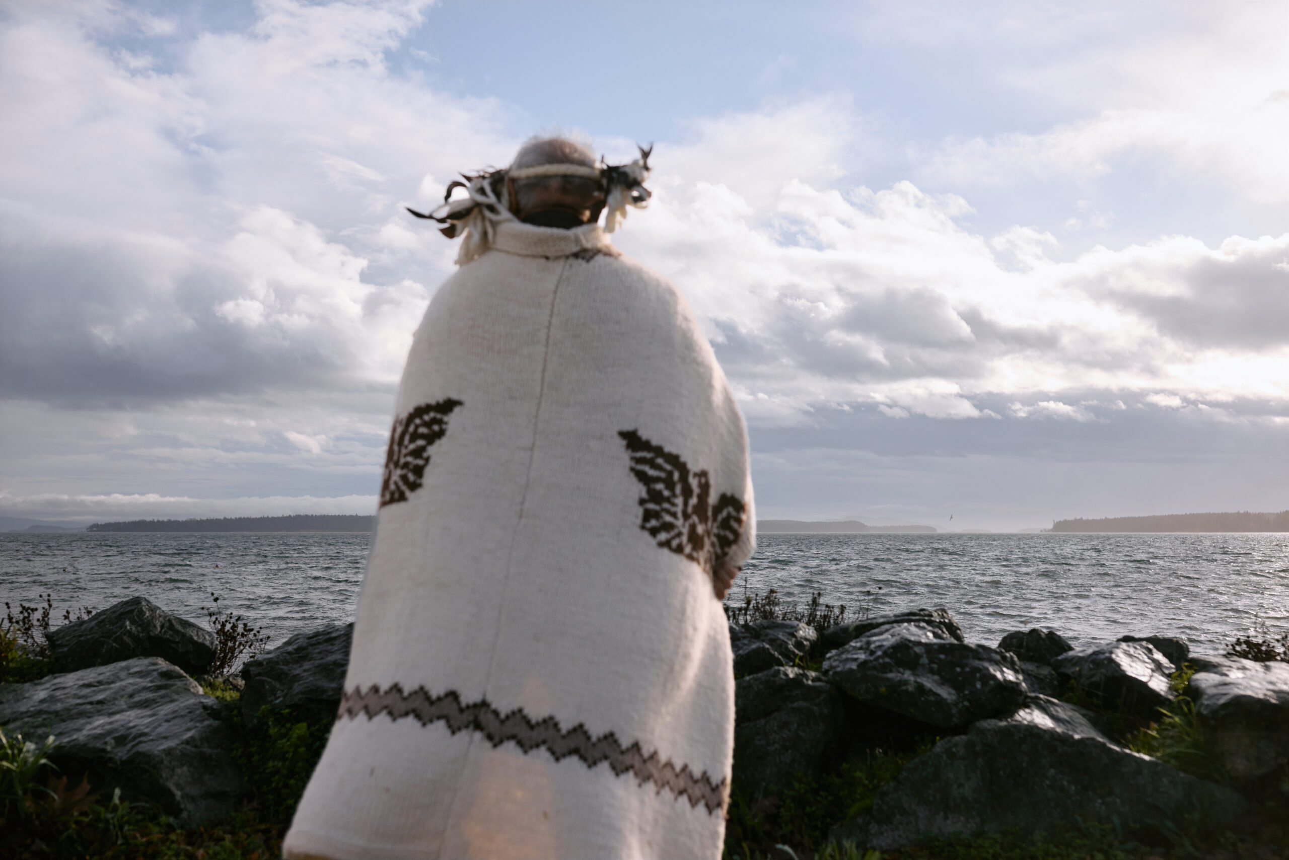 Chief Eric Pelkey faces the ocean, away from the camera. His wool regalia almost reaches the ground and is lightly lifted up by the wind, blowing slightly to the left. The sun pours in from the right. The same soft sunlight bathes the sky.