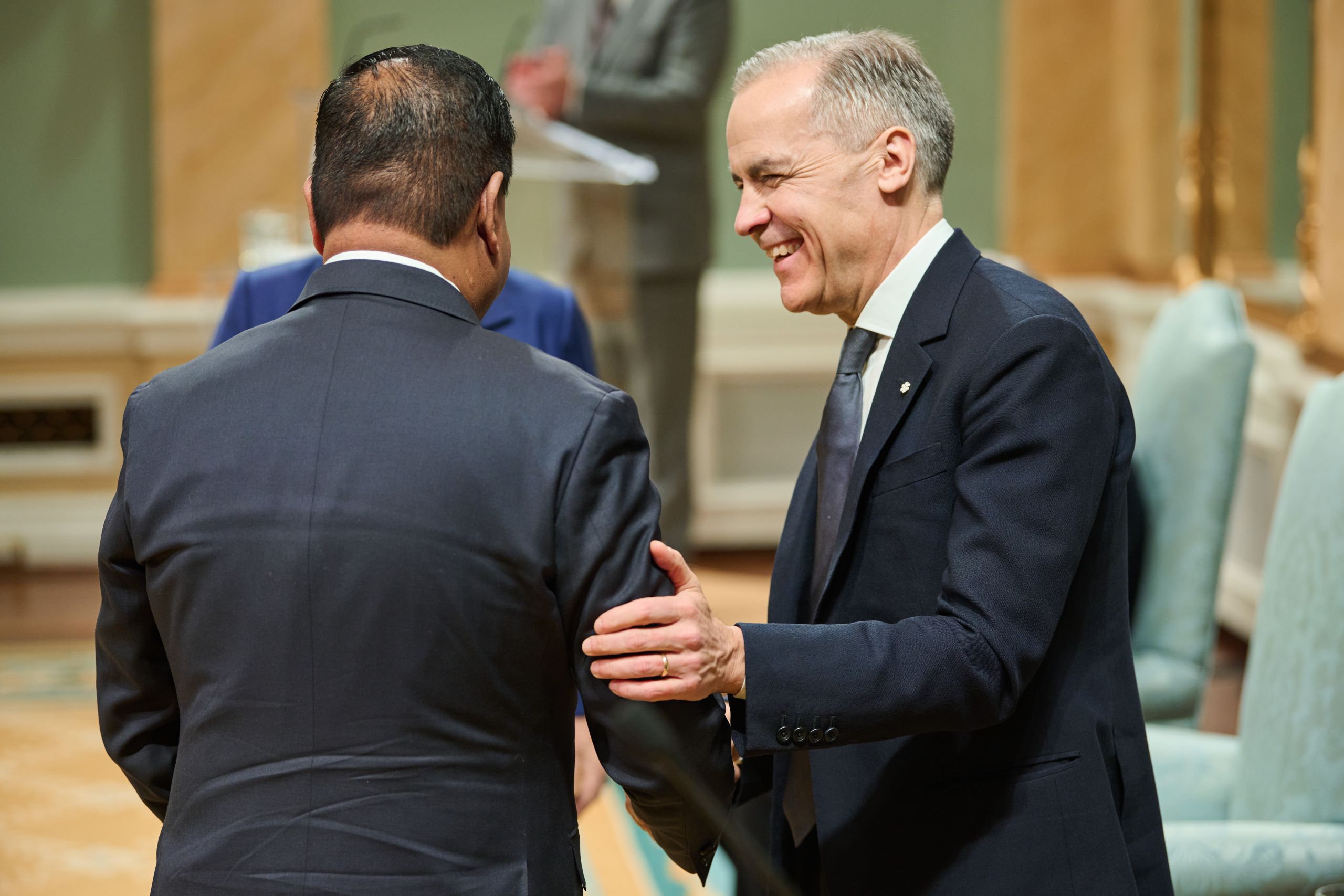 Man in a suit, right, smiles and grabs the arm of another man in a suit, who is facing away from the camera.
